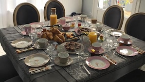 A dining table set with plates, cutlery, glasses, and a variety of food items, including pastries and a bottle of orange juice, in a room with large windows and a chandelier.