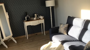 A room with a dark patterned wall, a blue sofa with cushions, a white lamp, a small table with a teapot, and a mirror on a stand.
