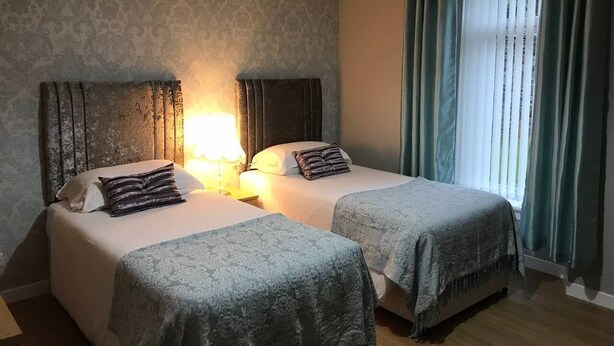 A hotel room with two beds, a nightstand with a lamp, a window with curtains, and a patterned wall. - 1 St Catherine’s City Cottage. Free Parking & WiFi (PERTH)