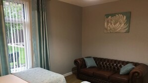 Ein Zimmer mit zwei Betten, einem braunen Ledersofa, einem Fenster mit Vorhängen und einem floralen Gemälde an der Wand.