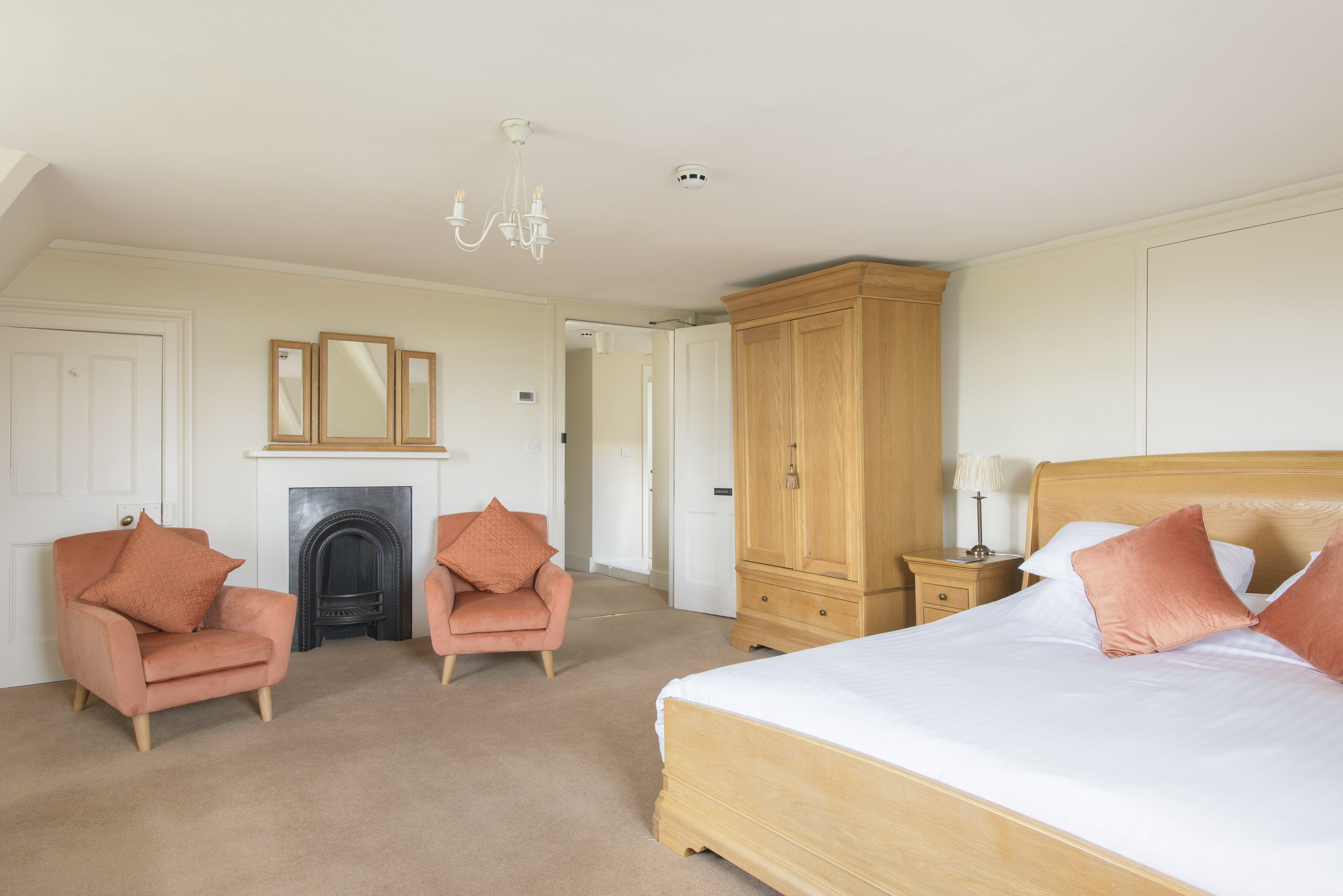 A bedroom with a bed, two armchairs, a wooden wardrobe, a nightstand, a lamp, and a fireplace.