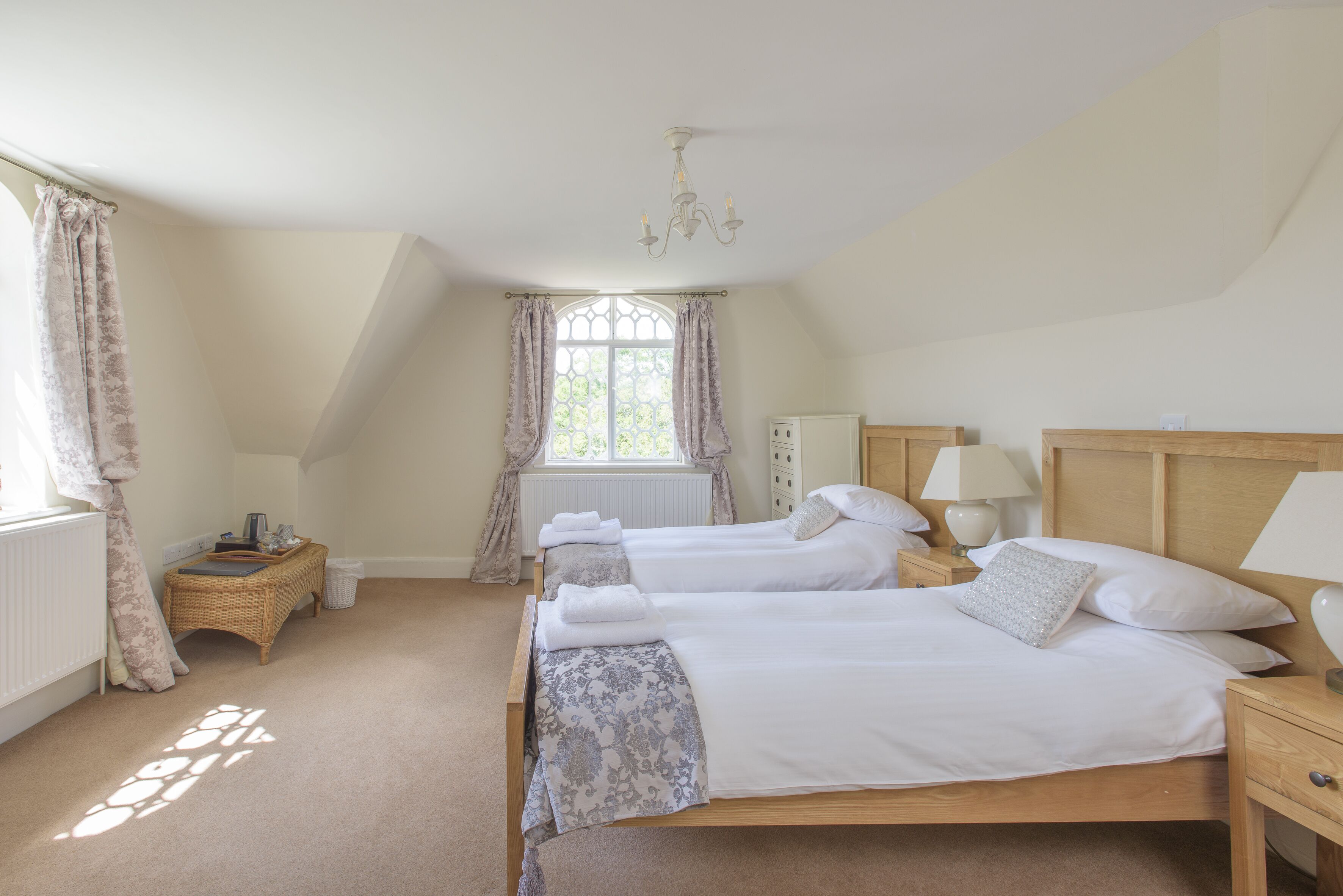 A bedroom with two single beds, a wooden headboard, bedside tables with lamps, a window with curtains, and a radiator.