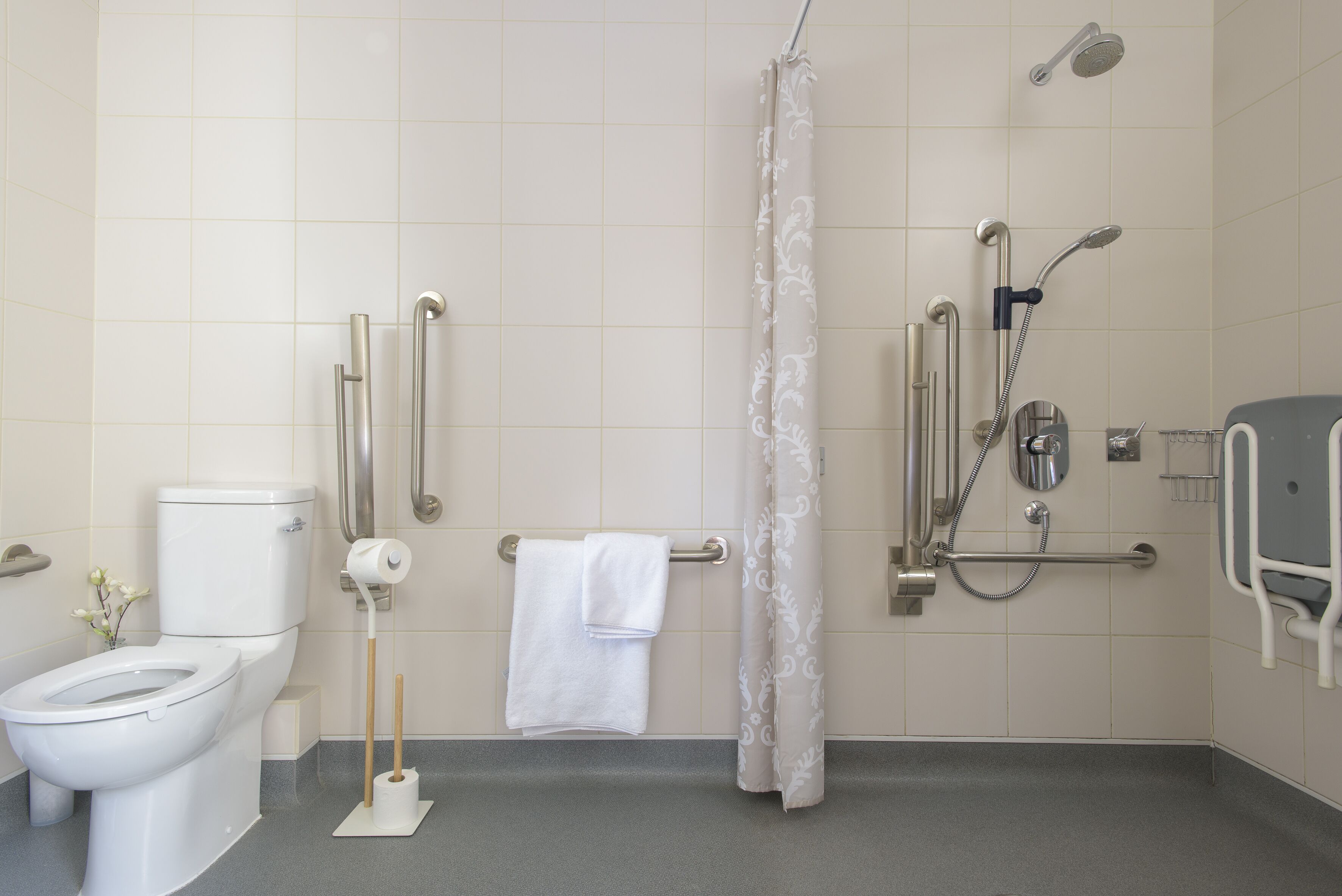 Ein Badezimmer mit Toilette, einer Dusche mit Haltegriffen, einer Haltestange in der Nähe des Waschbeckens und einem Handtuchhalter.