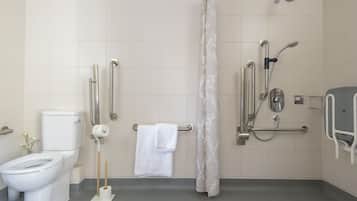 Ein Badezimmer mit Toilette, einer Dusche mit Haltegriffen, einer Haltestange in der Nähe des Waschbeckens und einem Handtuchhalter.