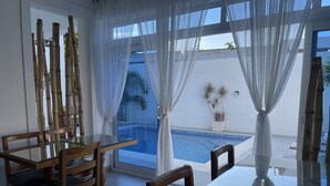 Una habitación con una mesa y sillas de vidrio, vistas a una piscina y decoración de bambú.