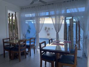 Un comedor con mesa de vidrio y sillas de madera, ventanas amplias con cortinas translúcidas y vista a una piscina y palmeras.