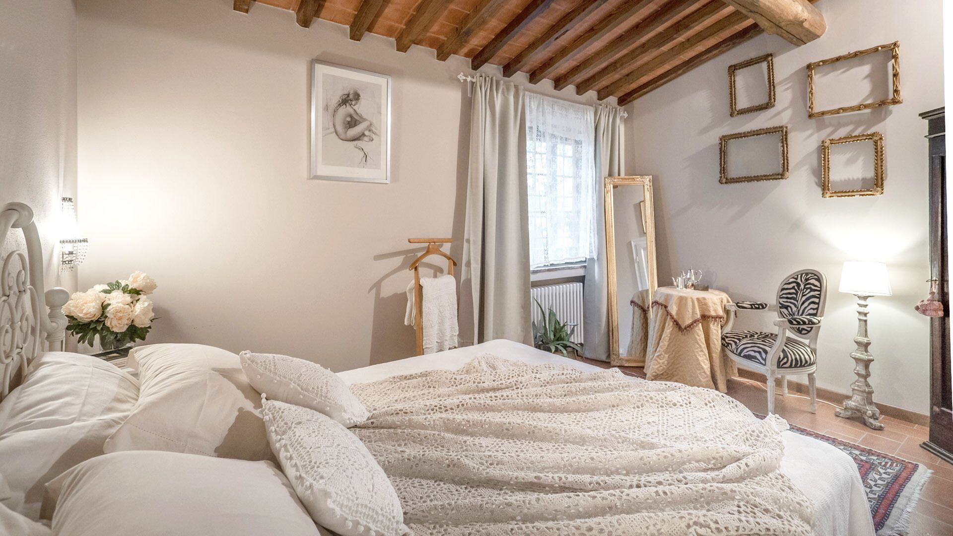 A bedroom with a large bed, a bedside table, a chair, a mirror, and a window with curtains.