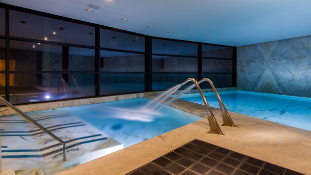 An indoor swimming pool with clear glass walls, a marble wall, and a metal ladder. - Encosta do Paraíso – Apartamentos Turísticos (Guimarães)