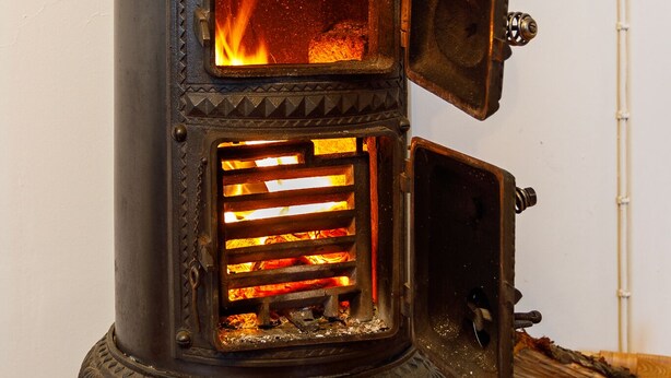 A wood-burning stove with a visible fire, a control dial, and a basket of firewood beside it. - Original North Country House (Laugar)