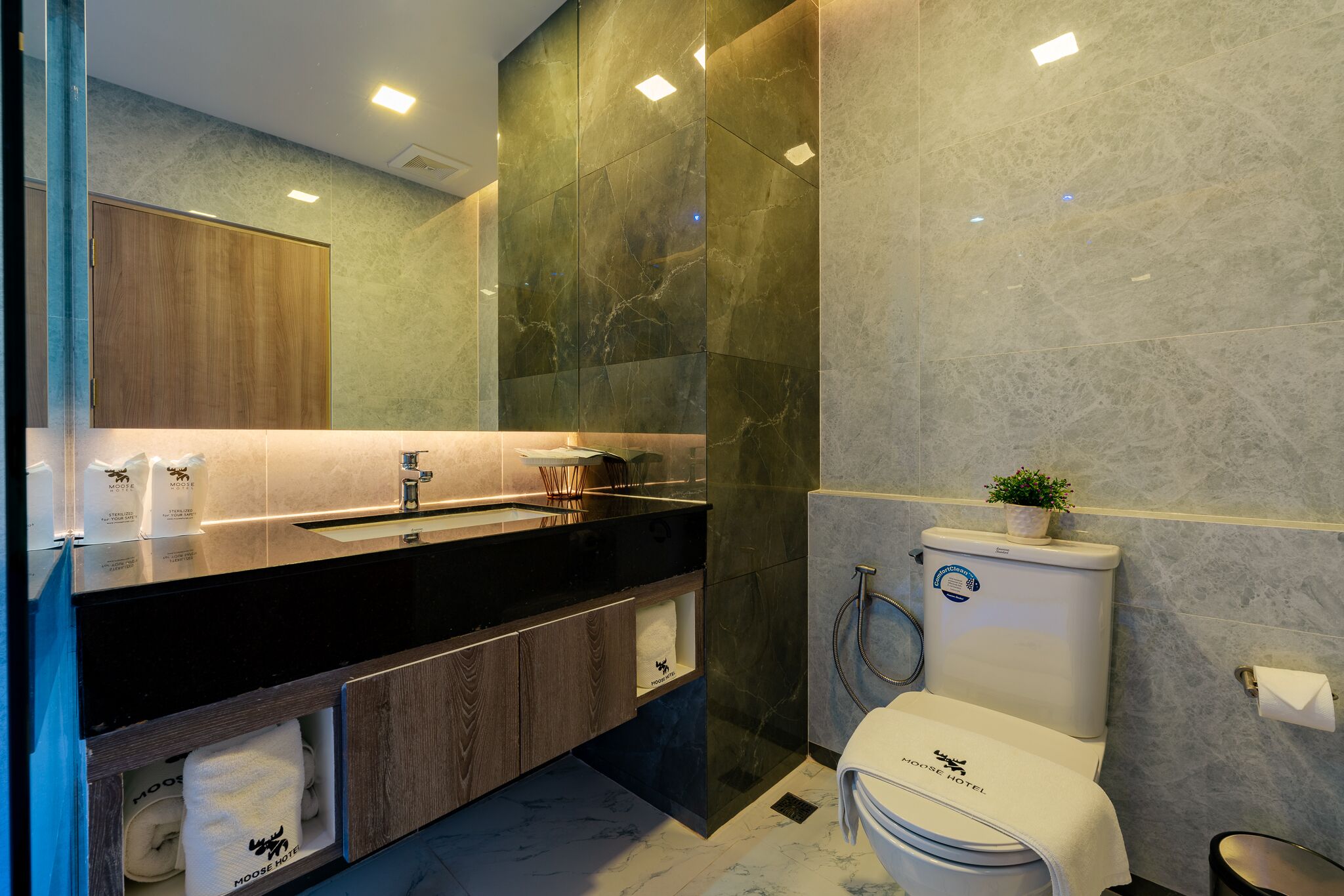 Un baño moderno con encimera negra, armarios de madera, pared de mármol y un inodoro con una toalla y una planta encima.