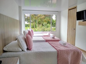 A bedroom with a bed, pillows, a TV, and a window with a view of trees. - Hotel Federman Park (Bogotá)