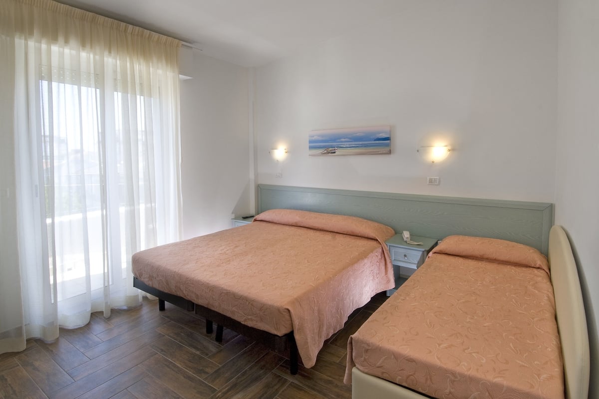 Quarto com duas camas, uma mesa de cabeceira, um quadro e uma janela com cortinas.