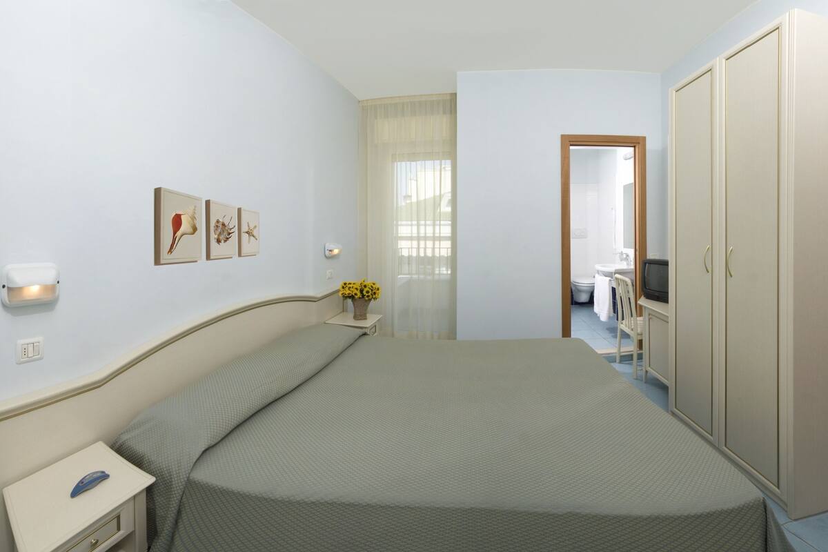 Quarto com cama, mesa de cabeceira, televisão, guarda-roupa e banheiro visível por uma porta aberta.