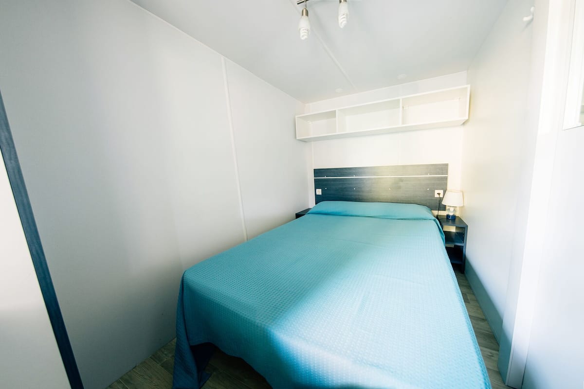 Ein kleines, ordentlich eingerichtetes Schlafzimmer mit einem Einzelbett, einem Nachttisch und einem Wandregal über dem Bett.