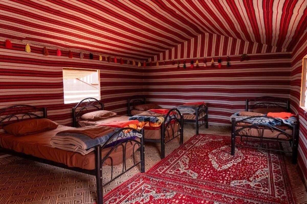 Une chambre avec quatre lits, chacun recouvert d’une couvre-lit à motifs différents. Un grand mur est percé d’une fenêtre, et le plafond de la chambre est rayé.