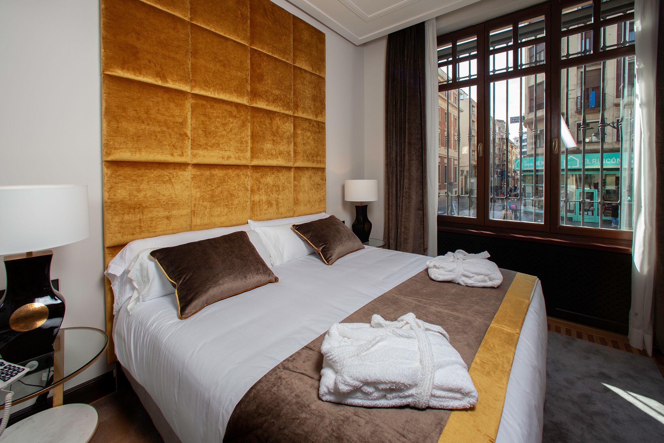 A hotel room with a large bed, a headboard, a window with curtains, and a bedside table with a lamp.