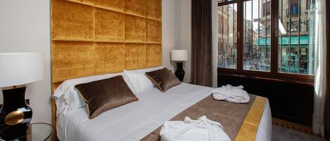 A hotel room with a large bed, a headboard, a window with curtains, and a bedside table with a lamp.