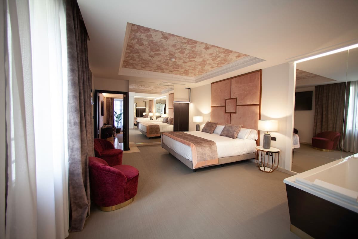 Une chambre d’hôtel avec un grand lit, un coin salon avec un fauteuil rouge, une petite table et un miroir.
