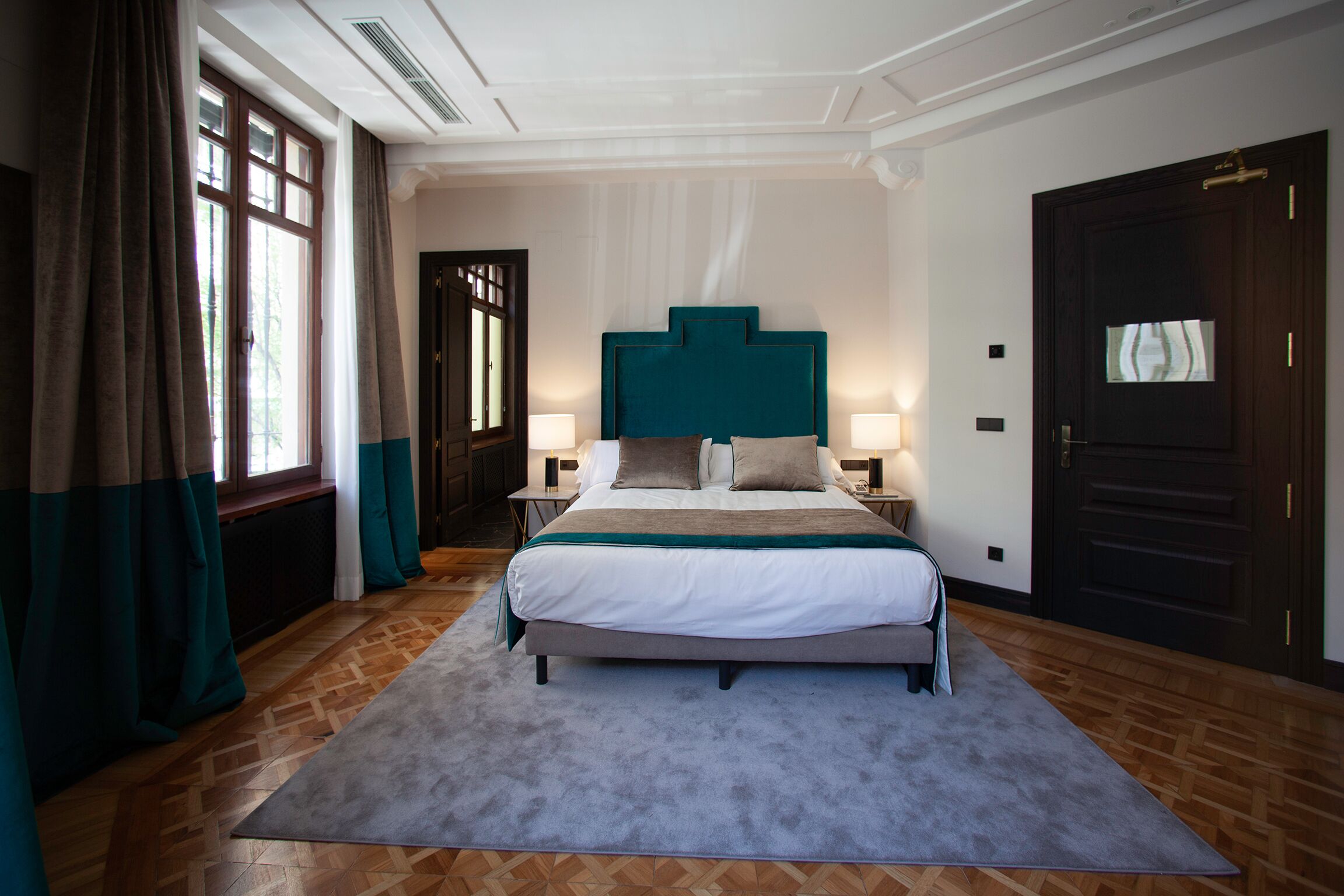 Een slaapkamer met een groot bed, twee nachtkastjes met lampen, een raam met gordijnen en een deur.