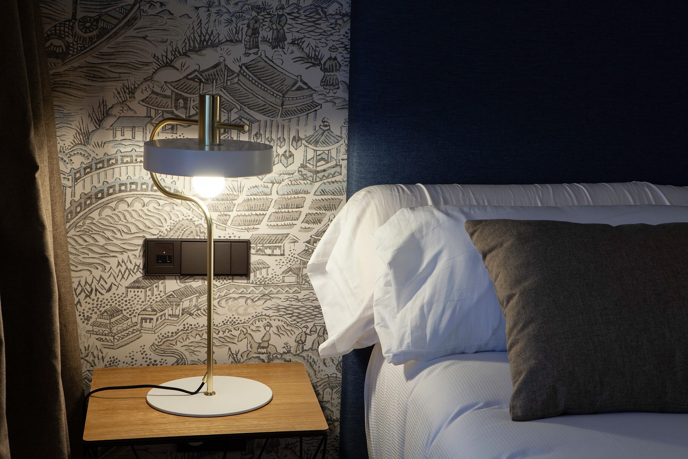 A neatly made bed with white and gray bedding, a bedside table with a lamp, and a wall with a detailed mural.