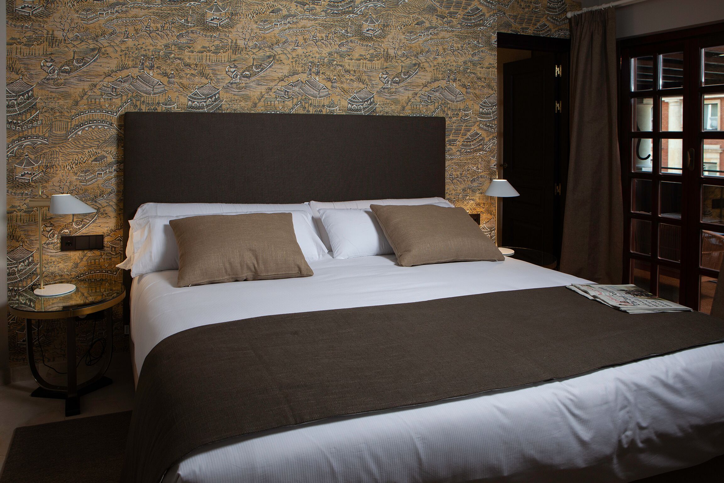 Una cama bien hecha con un cabecero oscuro, ropa de cama blanca y dos almohadas marrones. Hay dos lámparas de noche a cada lado de la cama.