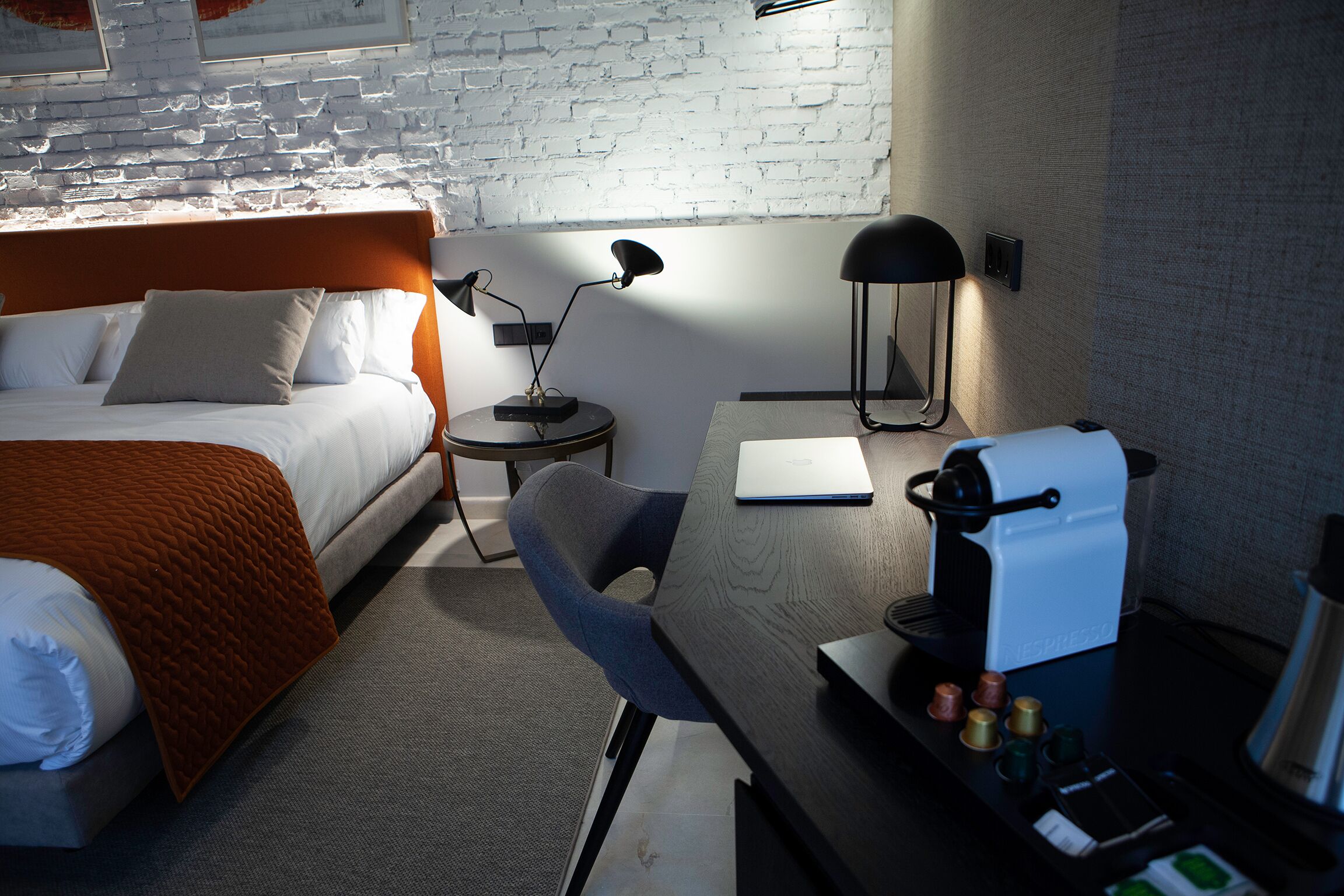 A hotel room with a bed, a desk with a coffee machine, a chair, and a lamp.
