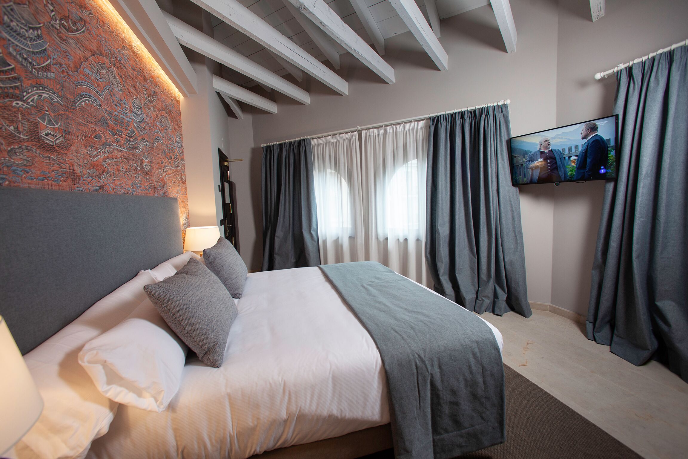 A modern hotel room with a large bed, a television mounted on the wall, and a patterned tapestry on the wall.