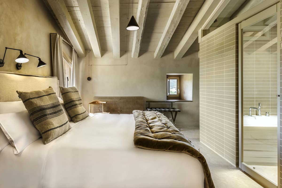 Quarto com cama grande, mesa de cabeceira, luminária e vista para o exterior por uma porta de correr.