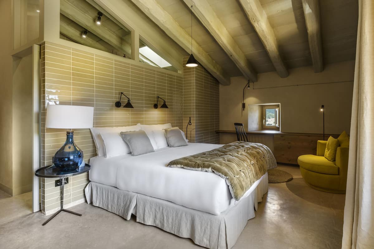 Um quarto espaçoso com uma cama grande, uma mesa de cabeceira com abajur e uma poltrona amarela.