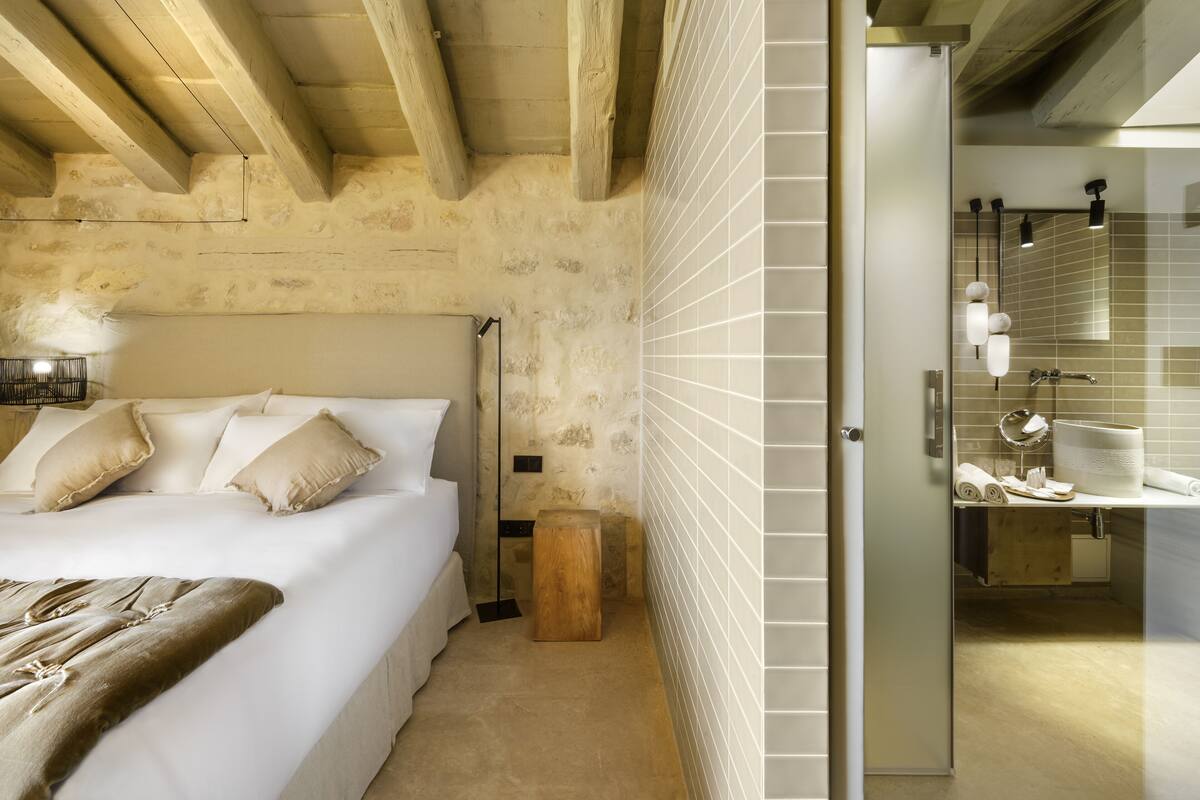 Quarto com cama grande, vigas de madeira, um banquinho pequeno e banheiro com pia e espelho.