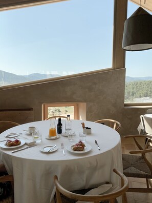 A round dining table set with plates, cutlery, glasses, and bottles, with a mountain view through a window.