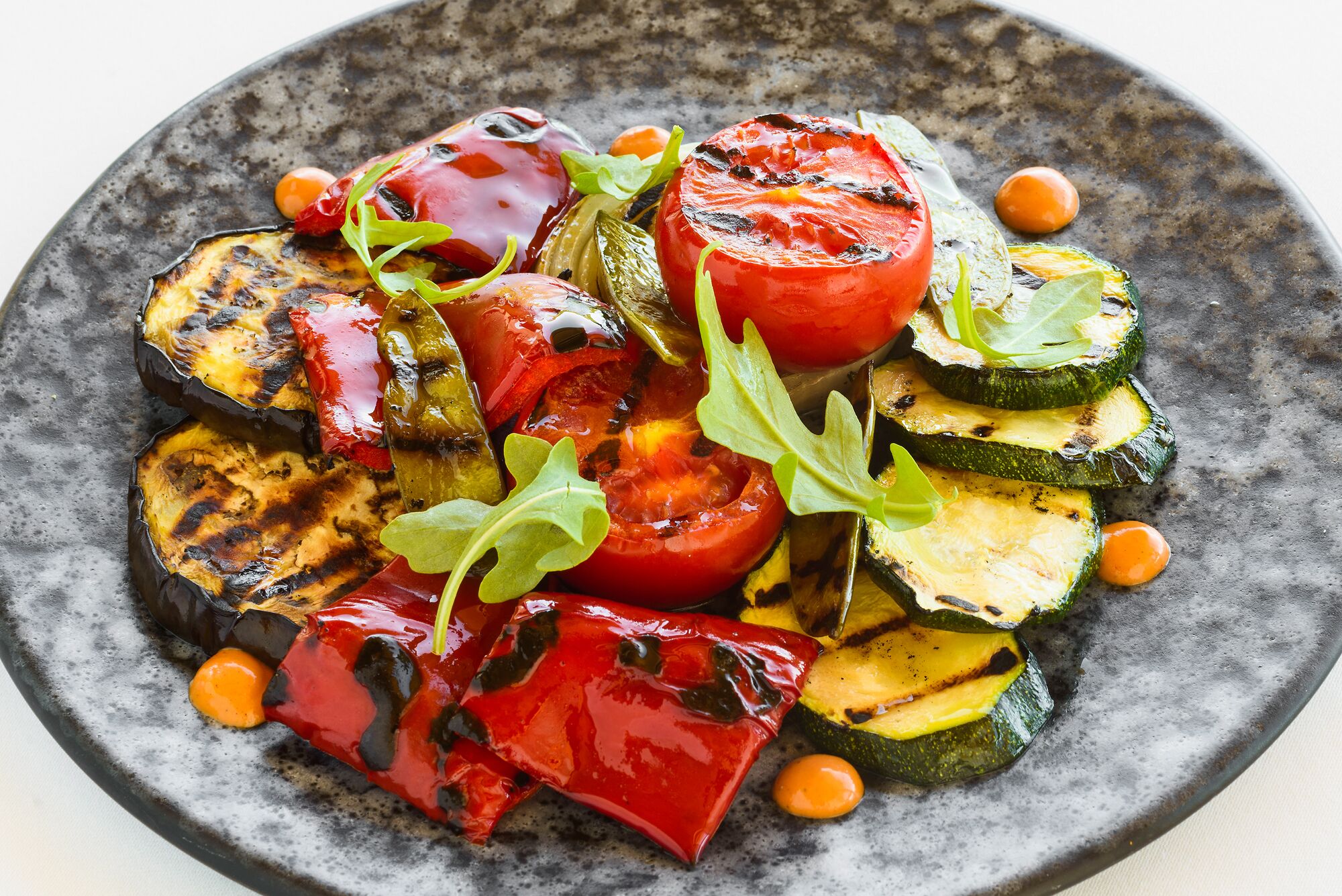 A plate of grilled vegetables including eggplant, zucchini, and tomatoes, garnished with greens and chickpeas.