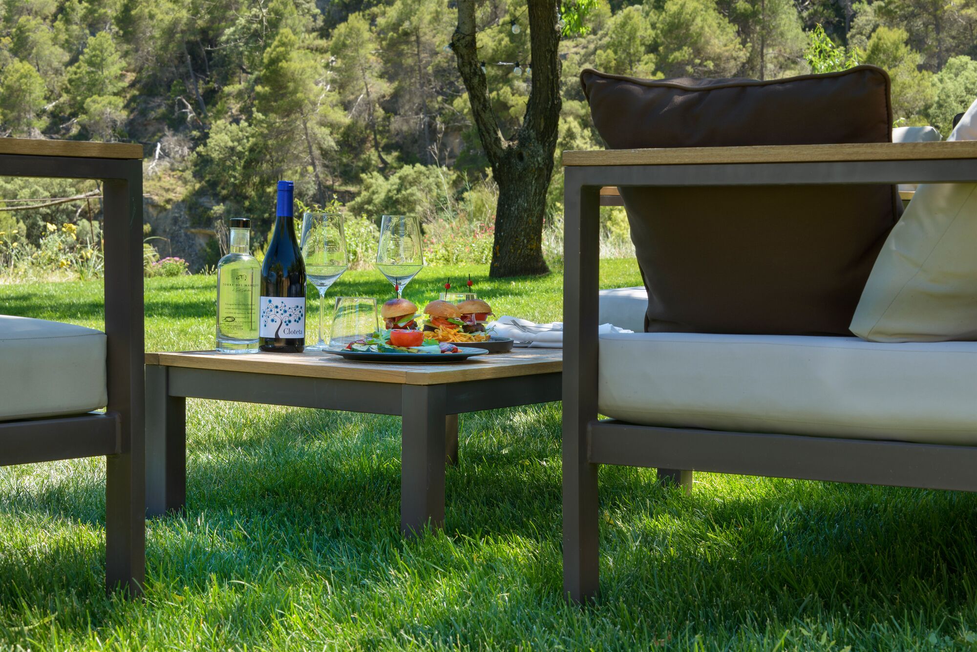 A picnic setup with a bottle of wine, glasses, and a plate of food on a table in a grassy outdoor area.
