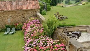 Un bâtiment en pierre avec un toit recouvert de tuiles, entouré d’une pelouse bien entretenue et d’une rangée de fleurs roses.