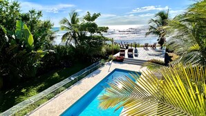 A swimming pool with a view of the ocean, surrounded by palm trees and a deck with chairs.