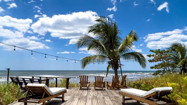 A wooden deck with lounge chairs and tables overlooking the ocean, palm trees, and string lights.