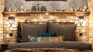 A cozy bedroom with a brick fireplace, a wooden shelf with decorative items, and a plush bed with multiple pillows. - Friendly Home - Special apartment "Old Bakery" (Dersdorf)