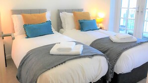 A hotel room with two beds, each with white bedding and blue and yellow pillows. There is a window with daylight coming through, and a lamp on the nightstand. - Seaside Cottage With Parking (Worthing)