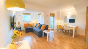 A modern living room with a sectional sofa, a dining area with a white table and yellow chairs, and a TV mounted on the wall. - Seaside Cottage With Parking (Worthing)