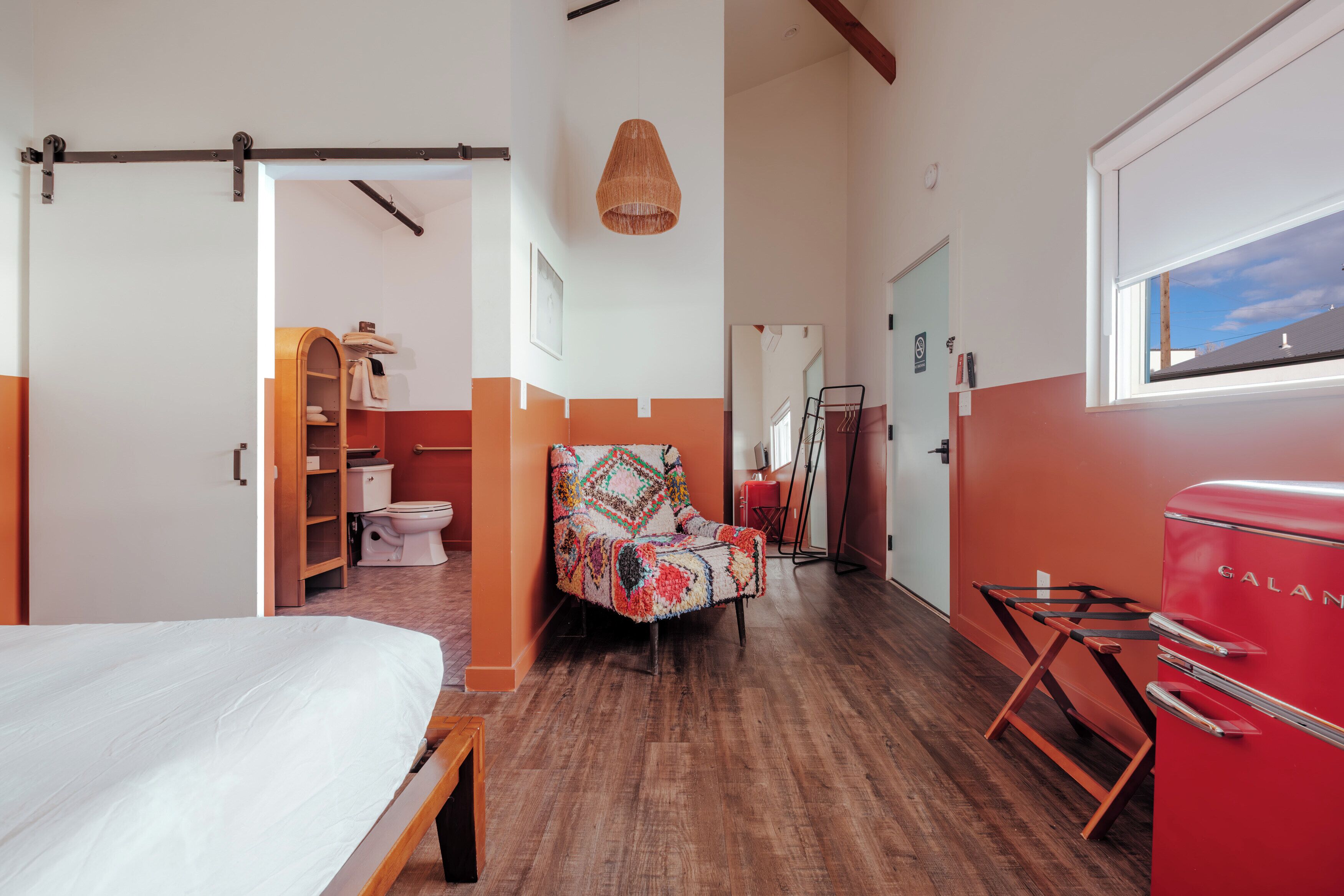A bedroom with a bed, a chair, a red appliance, and a hallway leading to a bathroom.