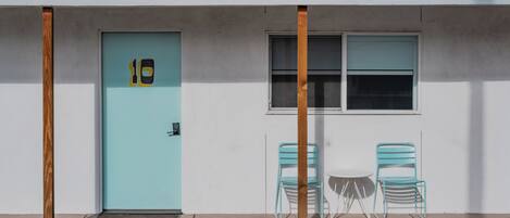Un edificio con una puerta azul claro, dos sillas de metal y una pequeña mesa.
