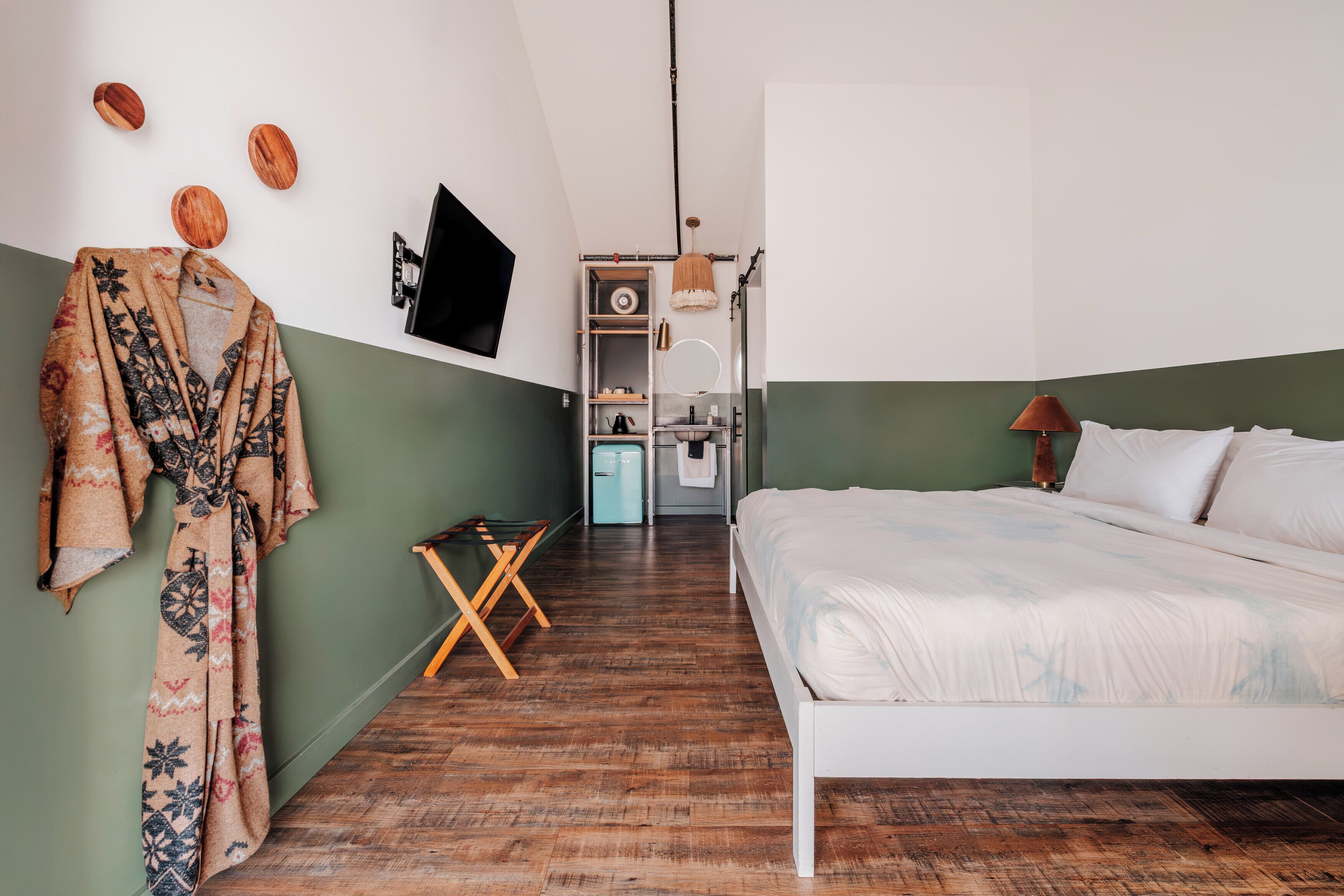 A bedroom with a bed, a wooden floor, a green wall, a robe hanging, a TV, a wardrobe, and a mirror.