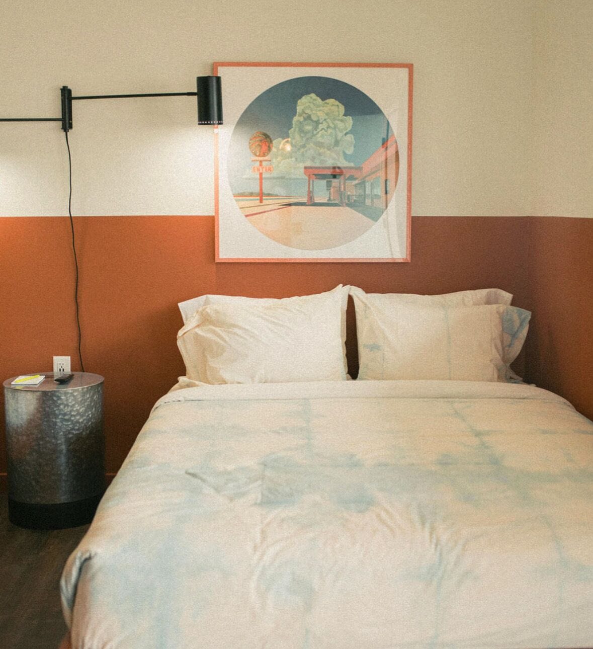 A bed with a white and blue patterned bedspread, a round side table with a remote control, and a framed picture on the wall.