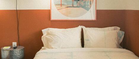 A bed with a white and blue patterned bedspread, a round side table with a remote control, and a framed picture on the wall.
