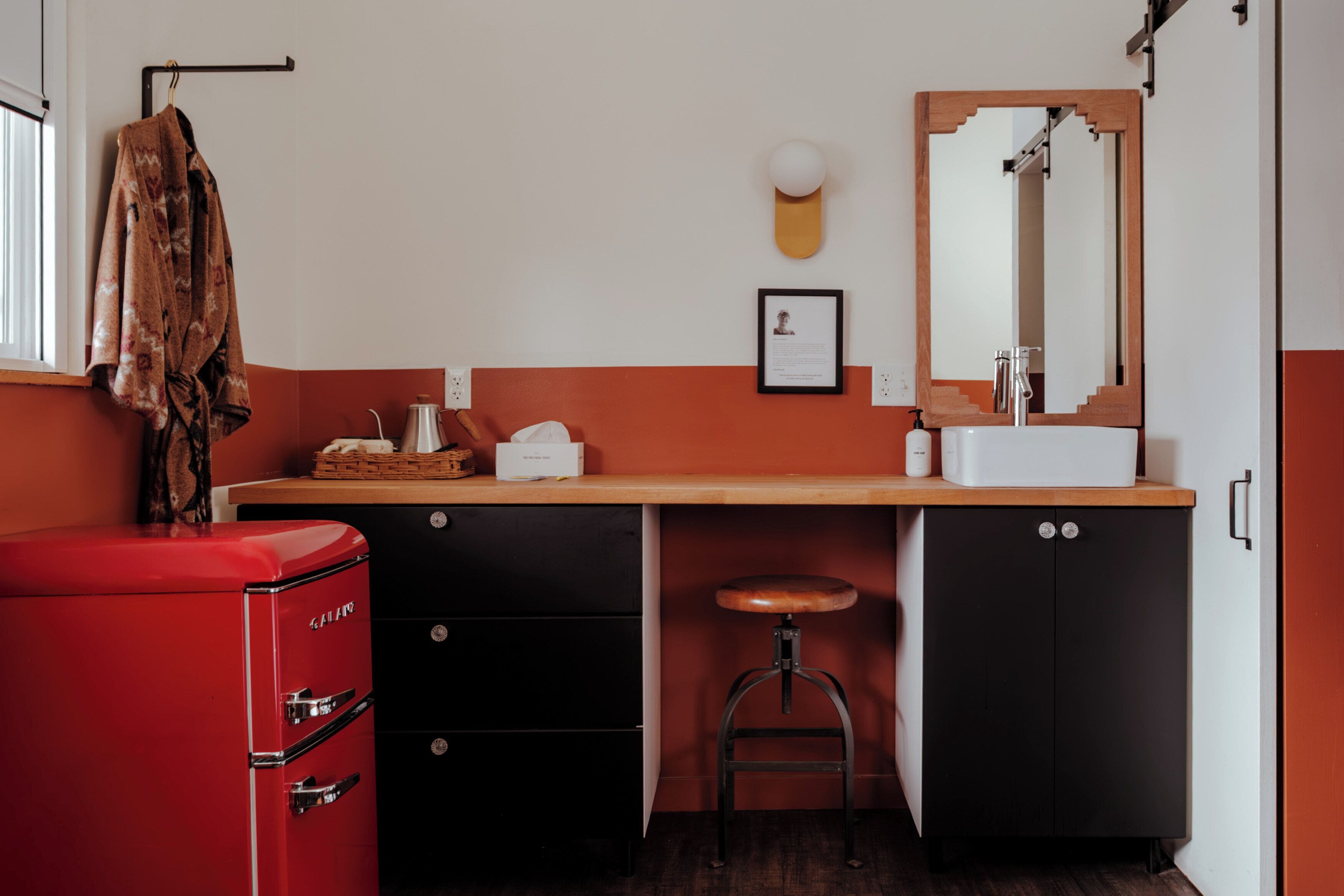 A compact kitchen with a red appliance, black cabinets, a wooden stool, and a mirror above the sink.