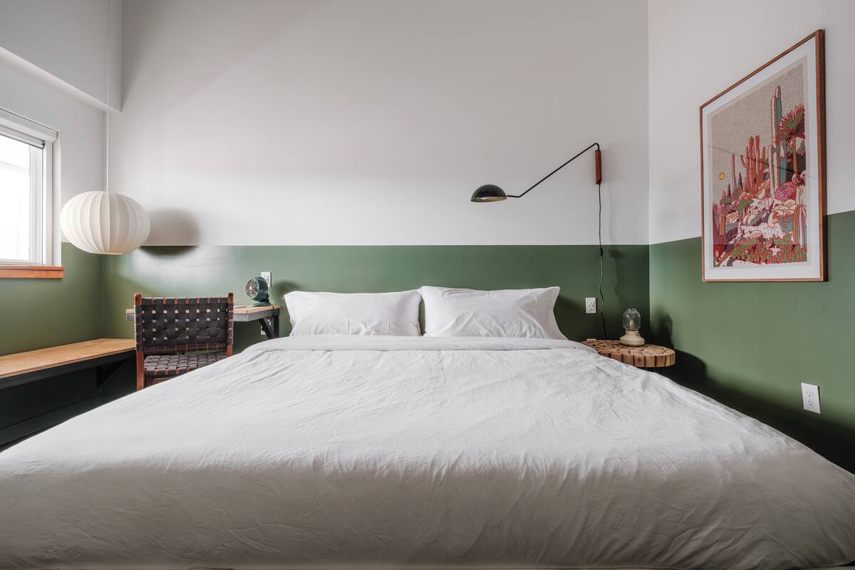 A neatly made bed in a room with a green accent wall, a desk with a chair, a lamp, and a framed artwork.