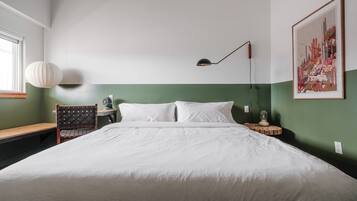 A neatly made bed in a room with a green accent wall, a desk with a chair, a lamp, and a framed artwork.
