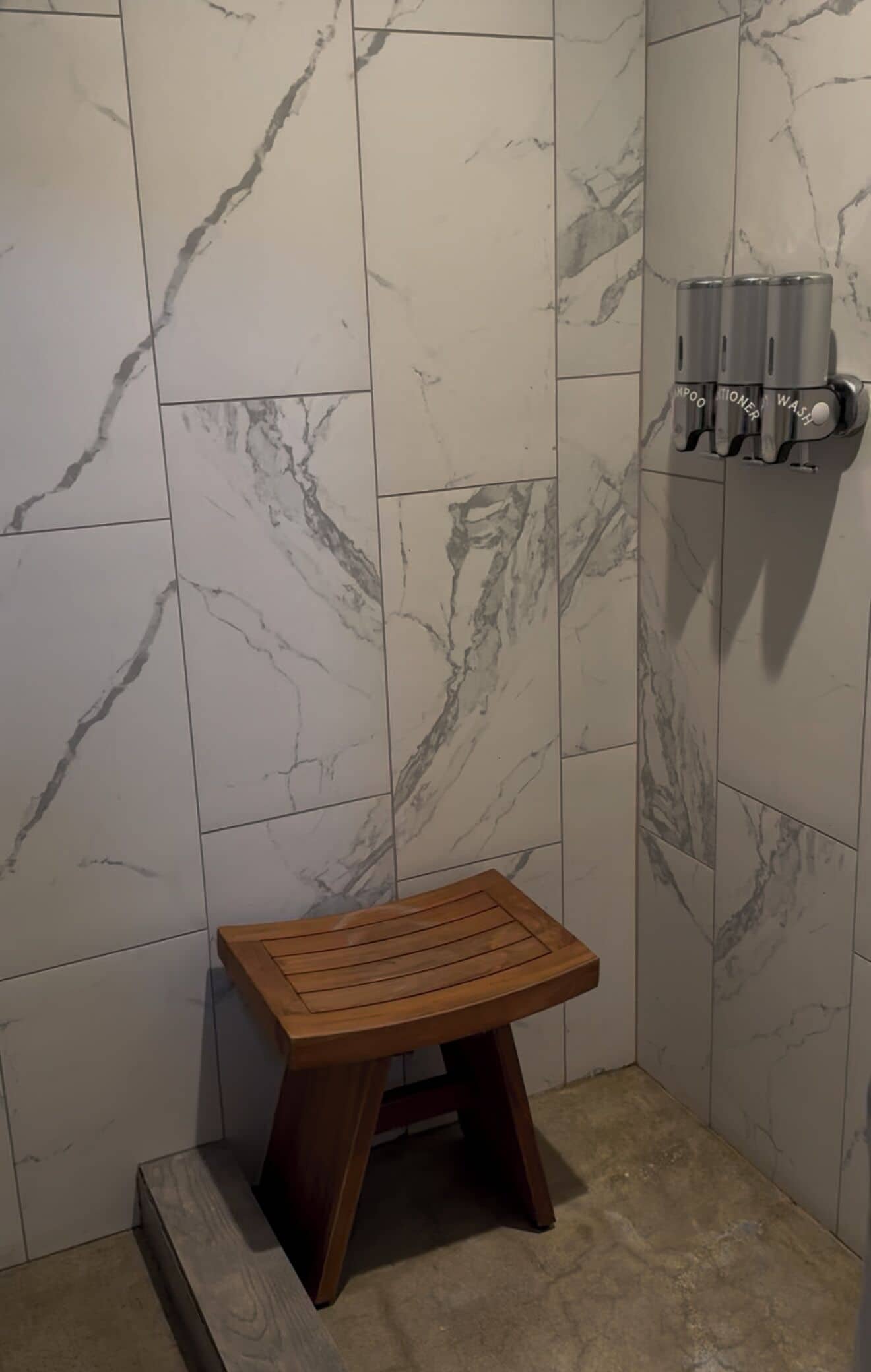 A bathroom with marble walls, a wooden stool, and a shampoo and conditioner dispenser.