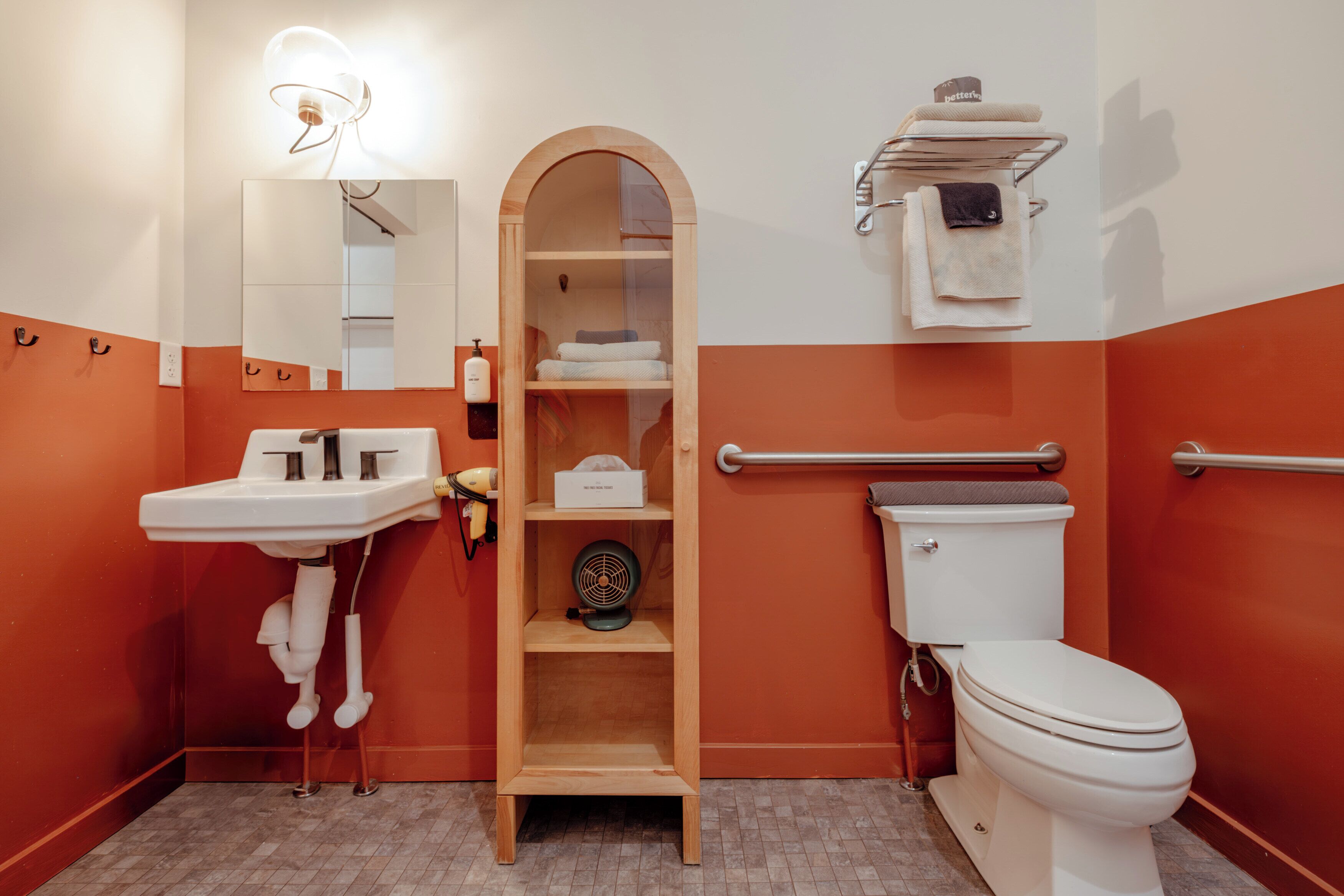 Un bany amb un lavabo, mirall, vàter i una estantària de fusta.