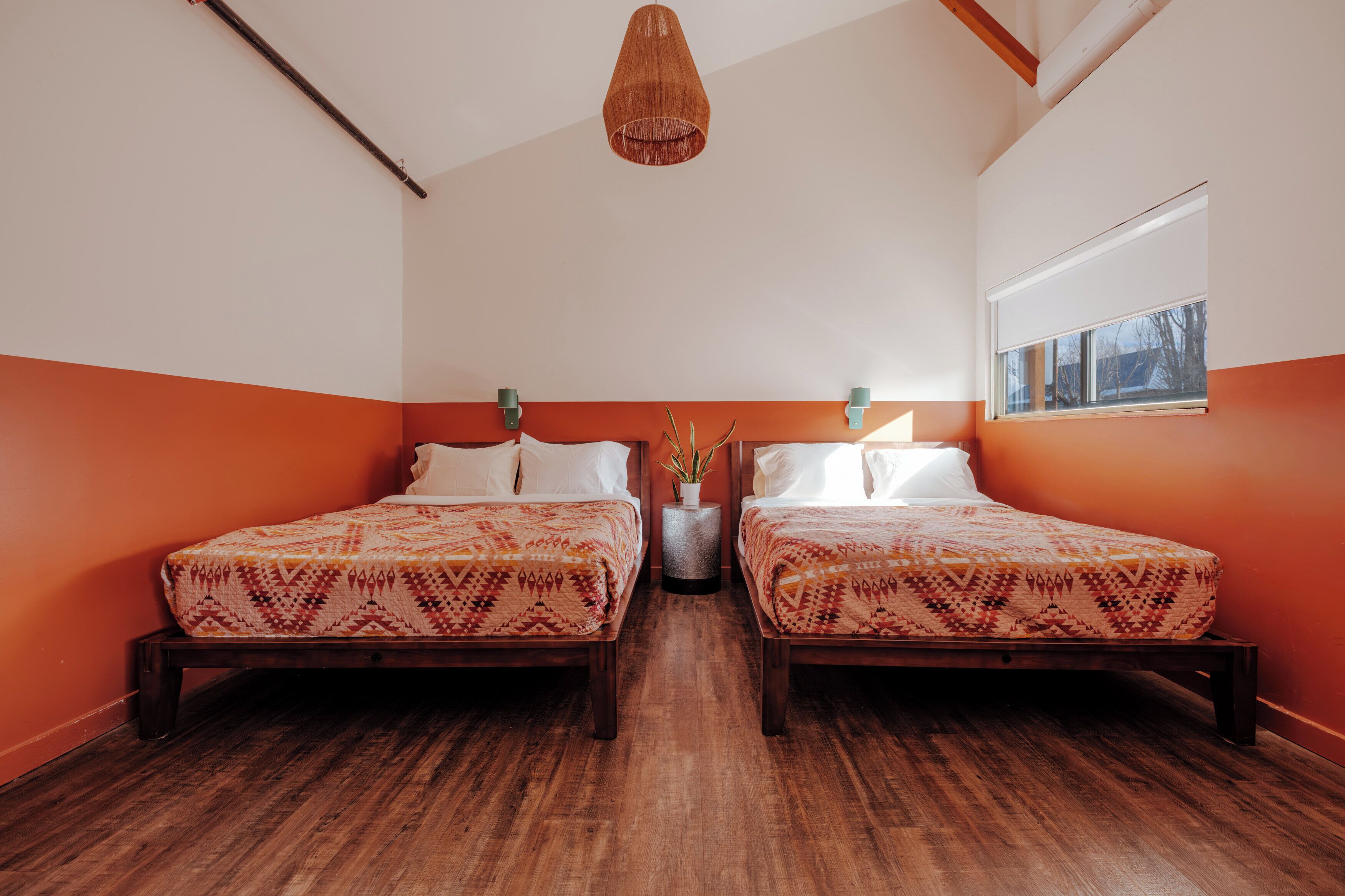 Une chambre avec deux lits, un luminaire suspendu, une fenêtre et une petite table d’appoint avec une plante.